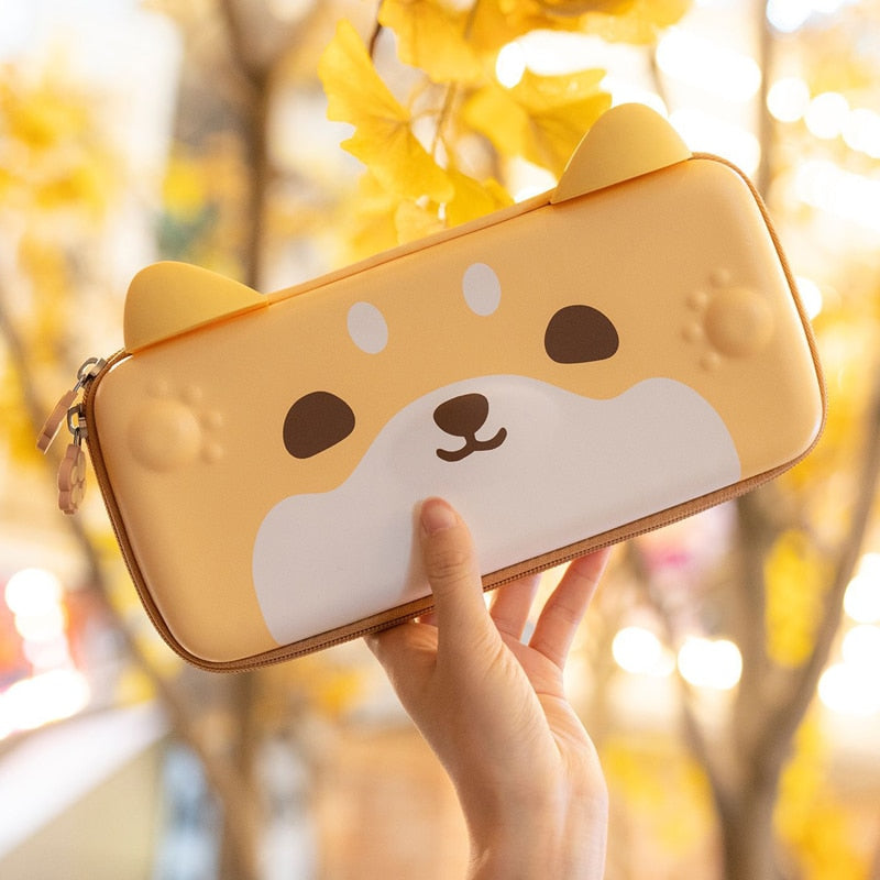 A hand holds a charming Shiba Pouch for Nintendo Switch by petimint, showcasing its tan hue with adorable bear-like ears and face design. The backdrop displays yellow autumn leaves on trees, setting a warm and cheerful ambiance—ideal for toting your essentials or your Nintendo Switch in stylish comfort.